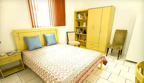 Suíte com cama de casal, armário amarelo, estante com livros e cadeira.