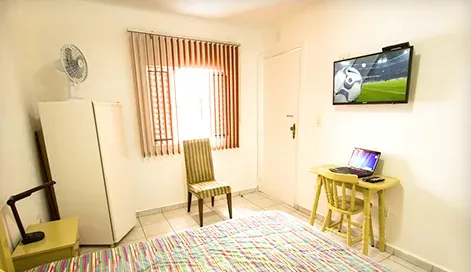 Suíte com TV LED na parede, geladeira, ventilador e escrivaninha com notebook.